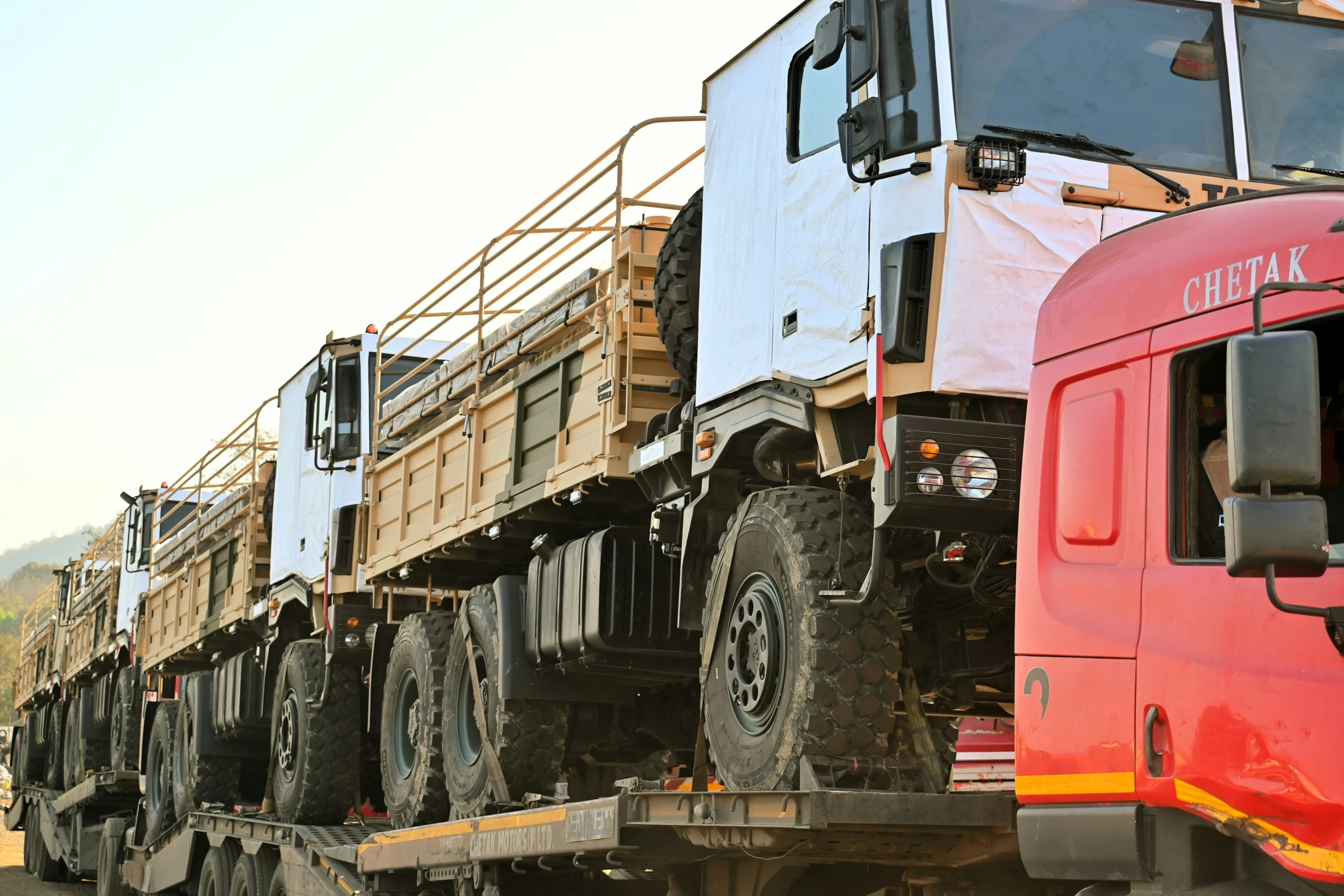 Military trucks in tan/beige color loaded on Chetak flatbed transport trailers, partially covered with white protective sheeting
